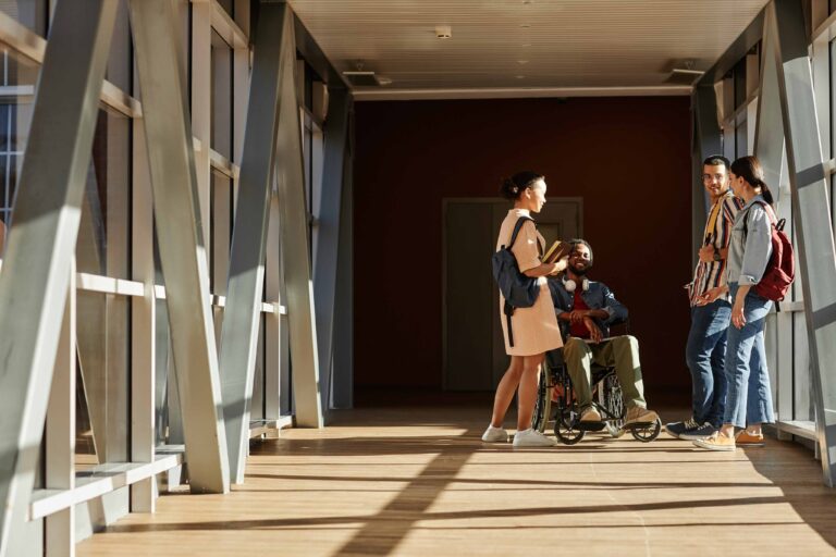 Four young adults, including one person in a wheelchair, are talking together in a sunlit hallway with large windows and backpacks.
