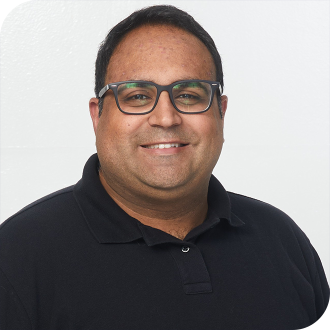 A man wearing glasses and a black polo shirt smiles at the camera against a plain white background.