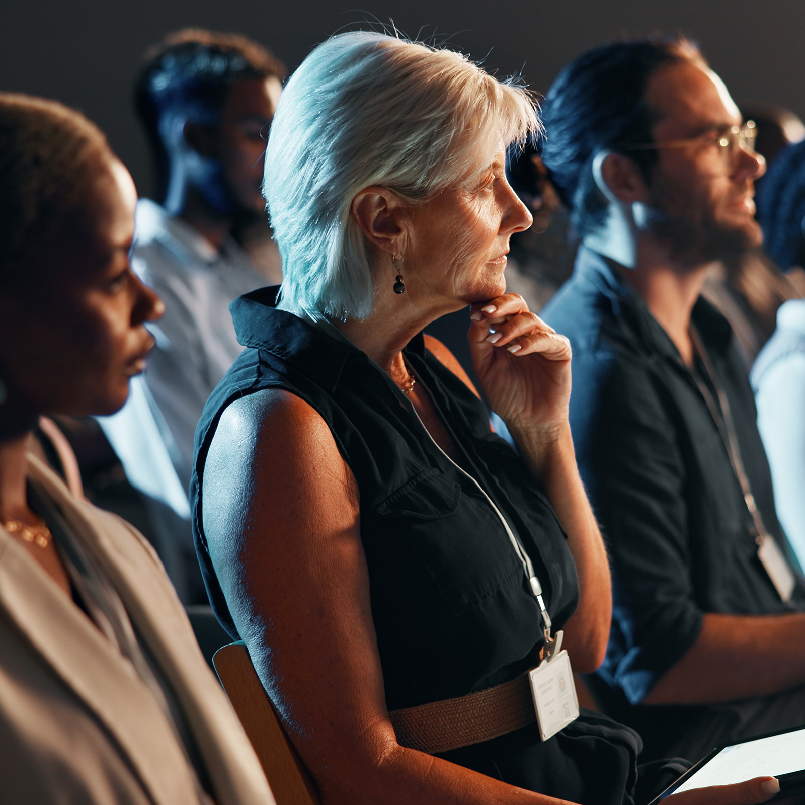 A group of people sit attentively in a row at an indoor event, wearing name badges and facing toward a speaker or presentation.