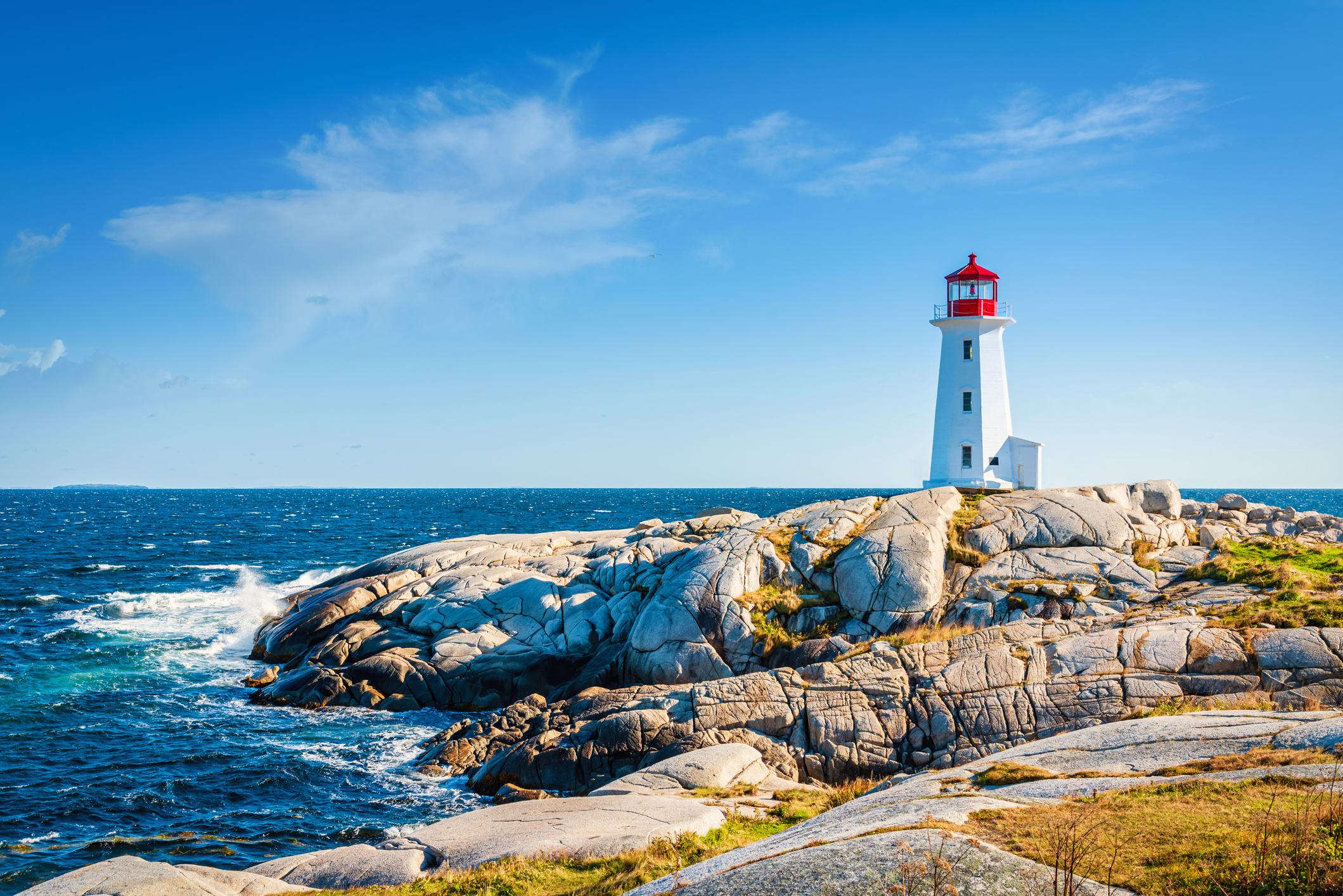 A white lighthouse with a red roof stands on a rocky coastline next to the Atlantic Ocean under a clear blue sky.