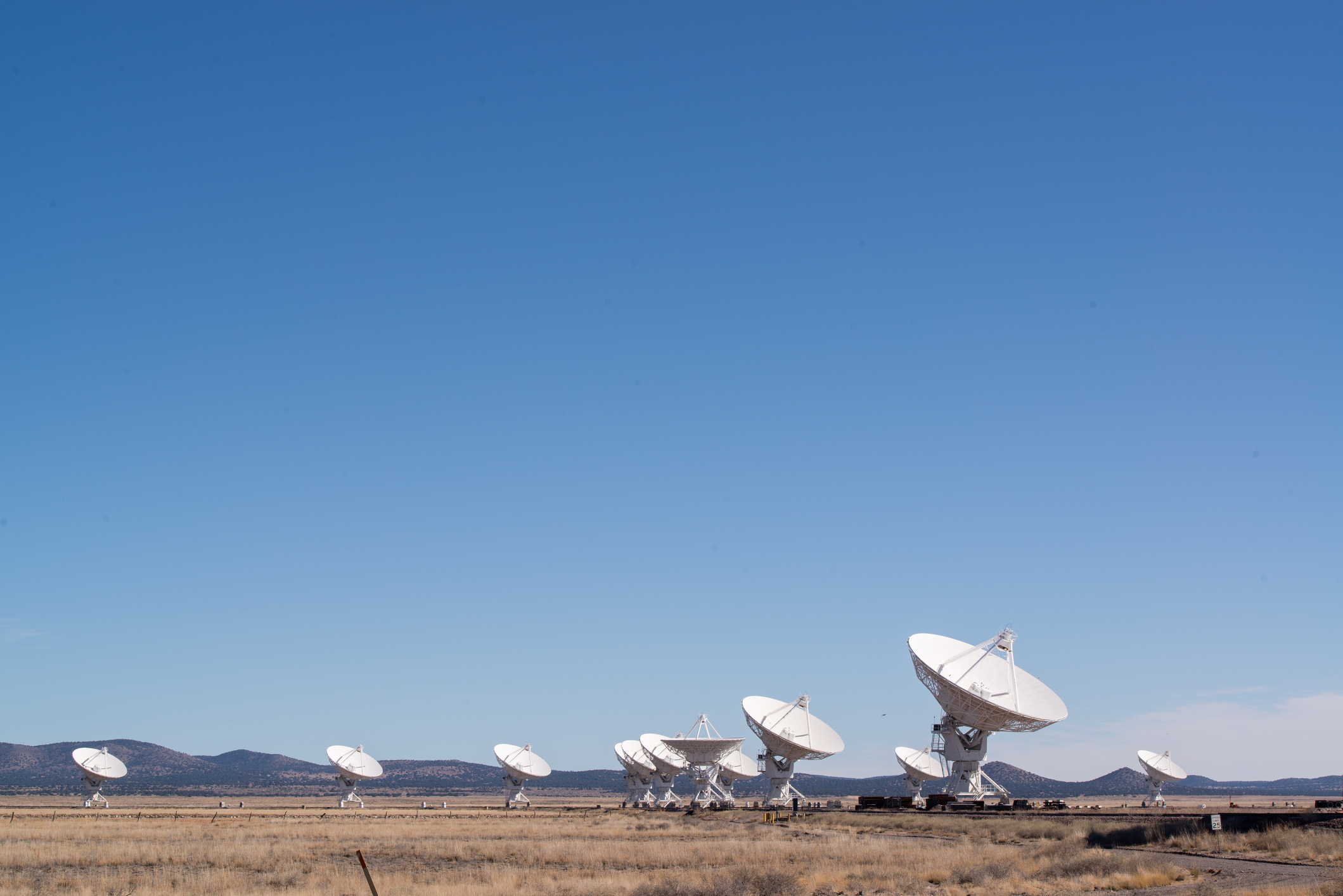 A row of large white radio telescopes stands in a dry field under a clear blue sky, with distant mountains in the background.
