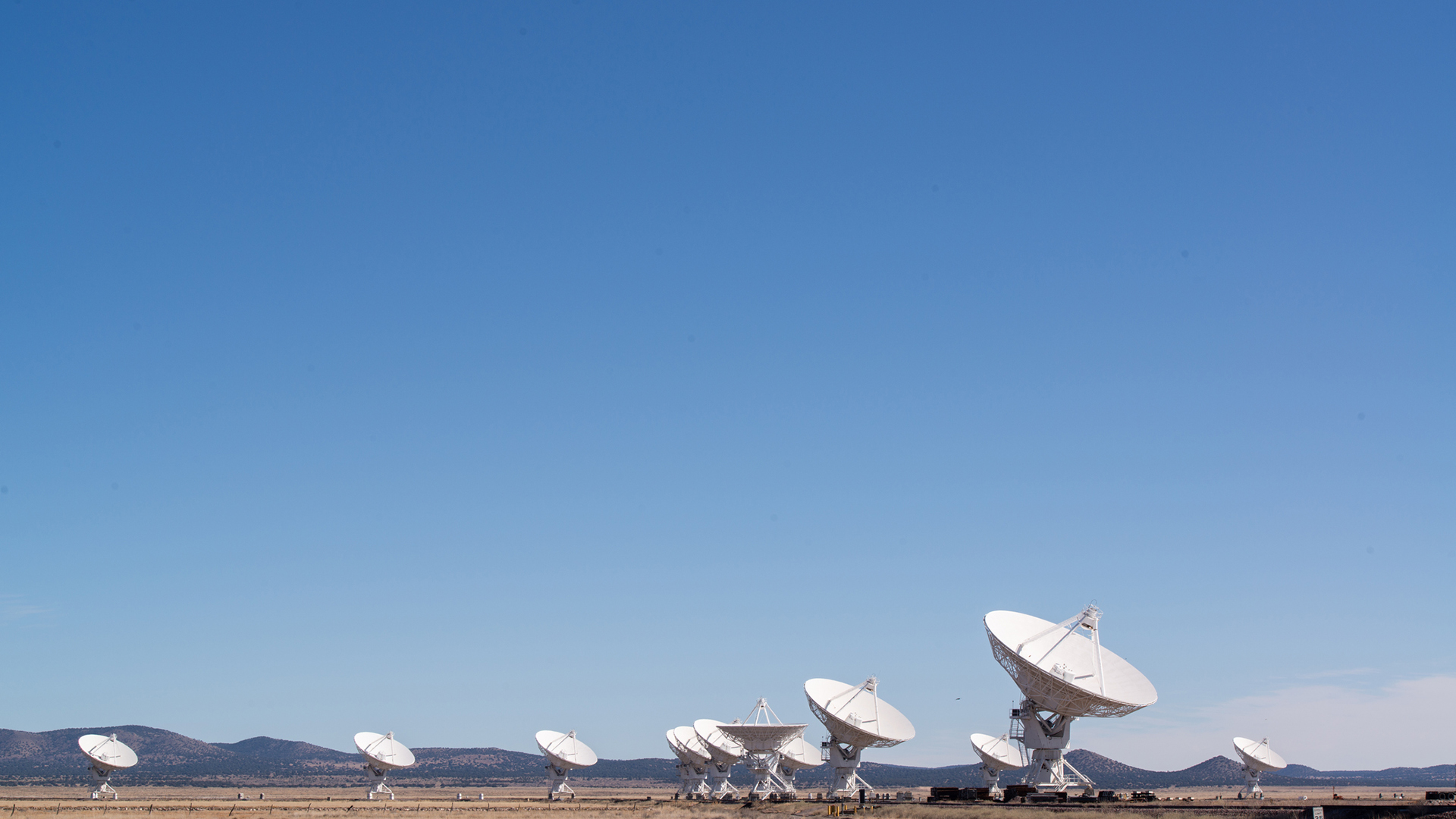 A row of large white satellite dishes sits on a flat landscape under a clear blue sky, with distant mountains visible in the background.