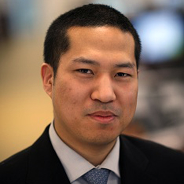 A man with short black hair, wearing a black suit, light blue shirt, and patterned tie, looks at the camera with a neutral expression. The background is blurred.